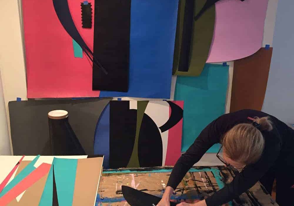 studio-beth-a-grillo-5A-Process-Paper A person works on a craft project in a studio with colorful abstract art pieces displayed on the wall.